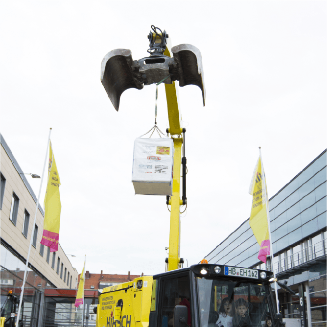 Ein gelber Lkw-Kran hebt mit einem großen Metallgreifer einen gefüllten Big Bag der Hirsch Gruppe vor einem Firmengebäude in die Höhe.