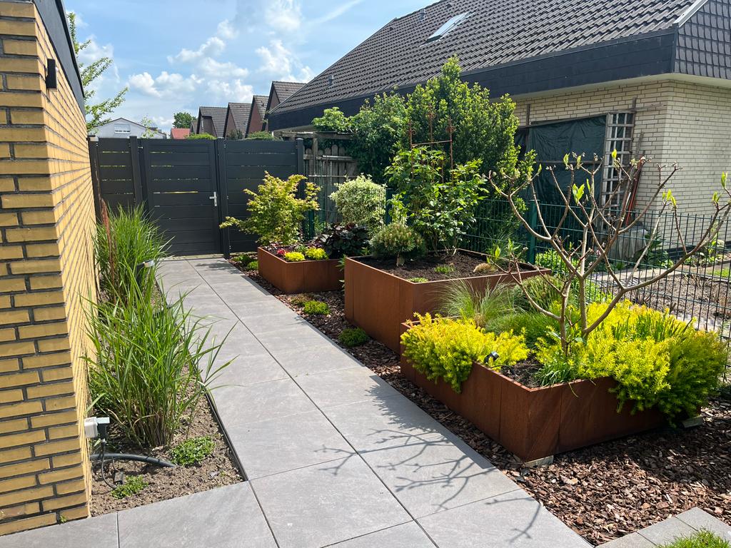 Ein modern angelegter Gartenweg aus großformatigen grauen Steinplatten, gesäumt von rostfarbenen Hochbeeten aus Cortenstahl und verschiedenen Zierpflanzen.