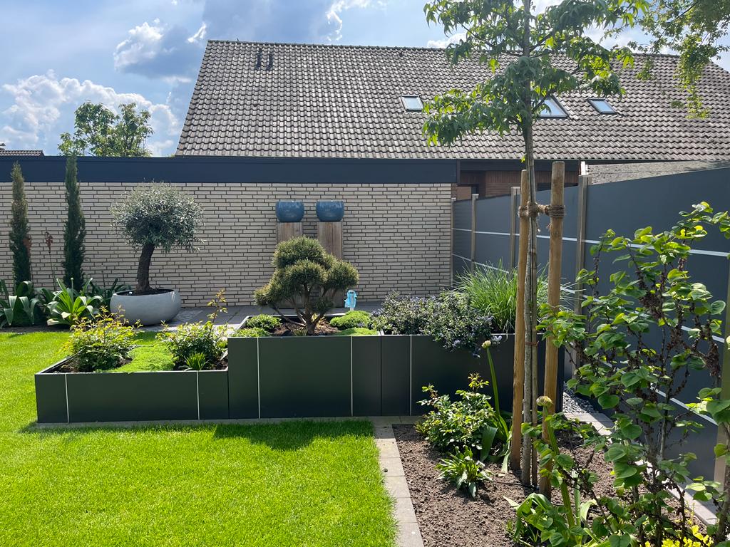 Gartenansicht mit Rasenfläche, Hecken, Bäumen und Sträuchern. Im Hintergrund eine Wand mit zwei blauen Töpfen und einem Dach mit Schindeln.