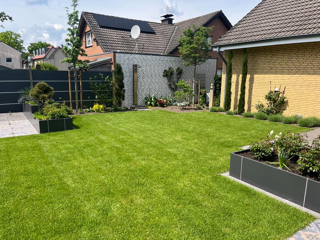 Gartenansicht mit gepflegter Rasenfläche, Hecken, Bäumen und Sträuchern. Im Hintergrund eine gelbliche gemauerte Hauswand.