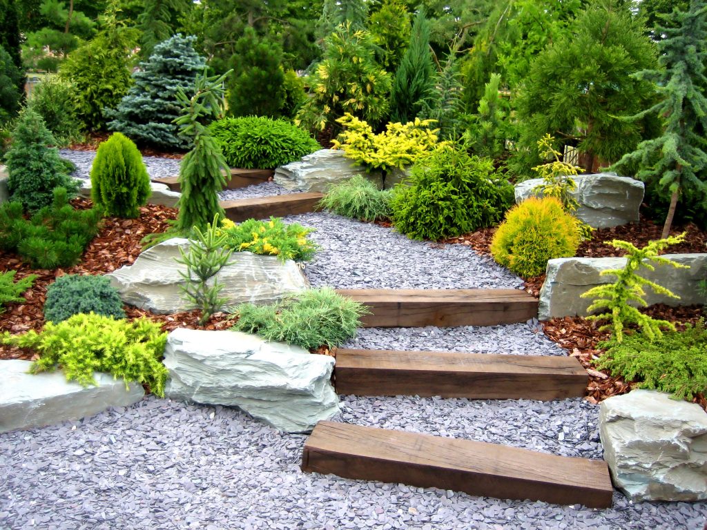 Ein aufwendig gestalteter Steingarten mit einem Weg aus hellem Splitt, rustikalen Holzstufen und großen Naturstein-Felsen, umgeben von verschiedenen Nadelgehölzen und Rindenmulch.