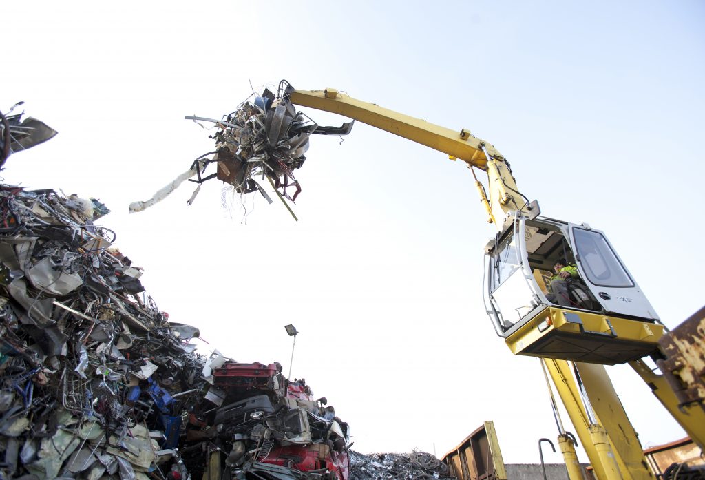 Ein gelber Hydraulikbagger greift mit einem Mehrschalengreifer eine Ladung Metallschrott von einem großen Haufen.