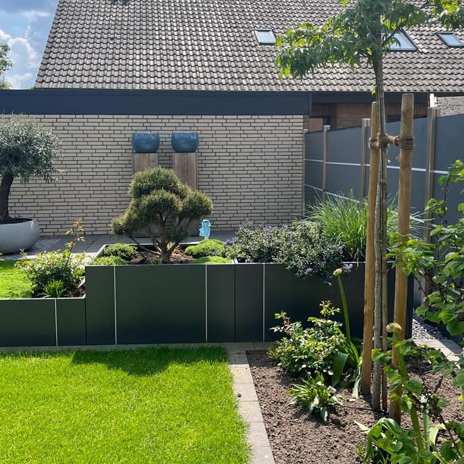 Gartenansicht mit Rasenfläche, Hecken, Bäumen und Sträuchern. Im Hintergrund eine Wand mit zwei blauen Töpfen und einem Dach mit Schindeln.