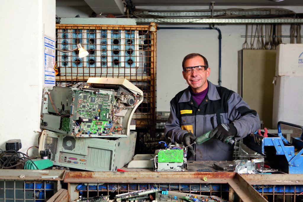 Ein lächelnder Mitarbeiter der Hirsch Recycling GmbH mit Schutzbrille demontiert fachgerecht einen PC an einem voll ausgestatteten Werkstattplatz.